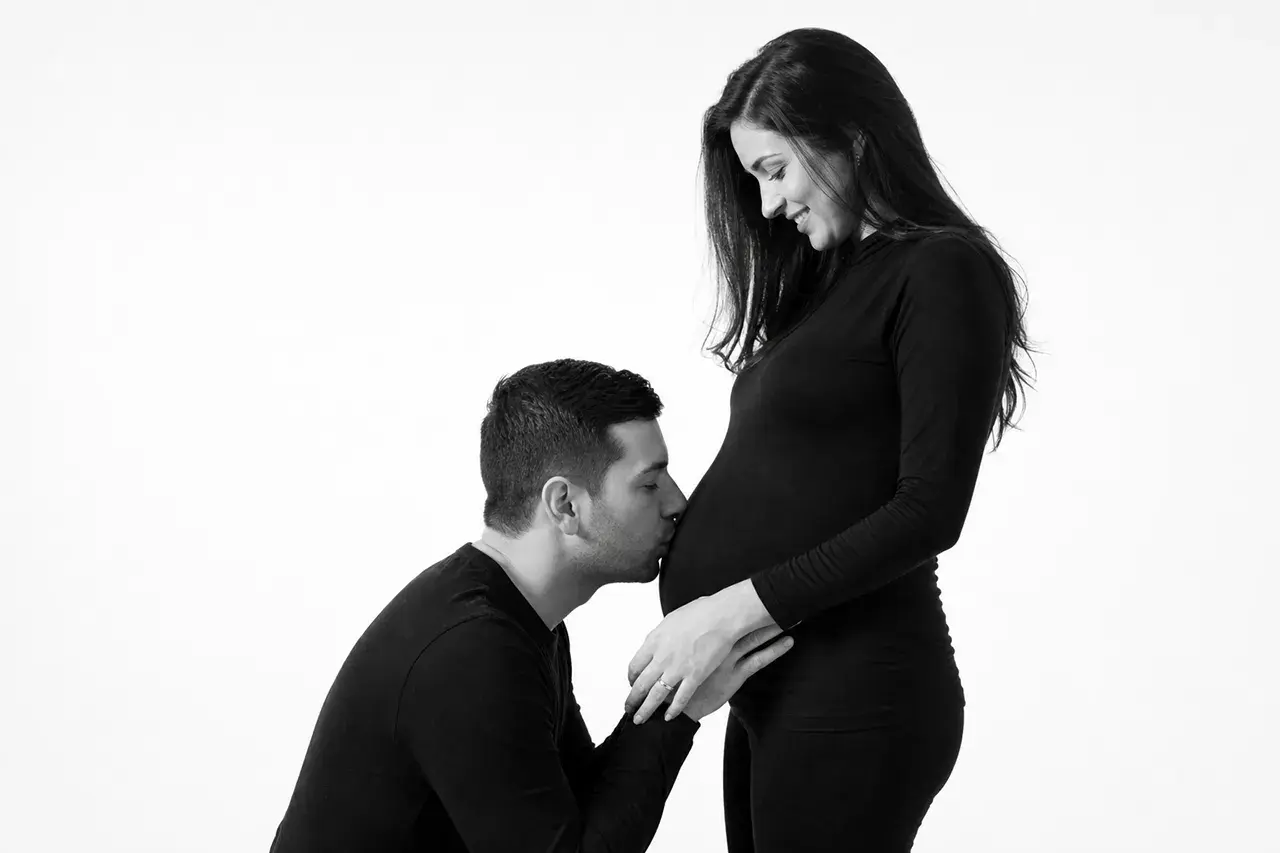Babybauchshooting in München: So bereitest du deine Studiofotos vor 3 Partner küsst den Babybauch einer schwangeren Frau, Schwarzweißaufnahme im Fotostudio